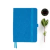 Aqua blue fabric-covered notebook with rounded corners, matching elastic band, pen loop, and dual ribbon bookmarks, shown with a green leafy branch and a floral 'joie' logo