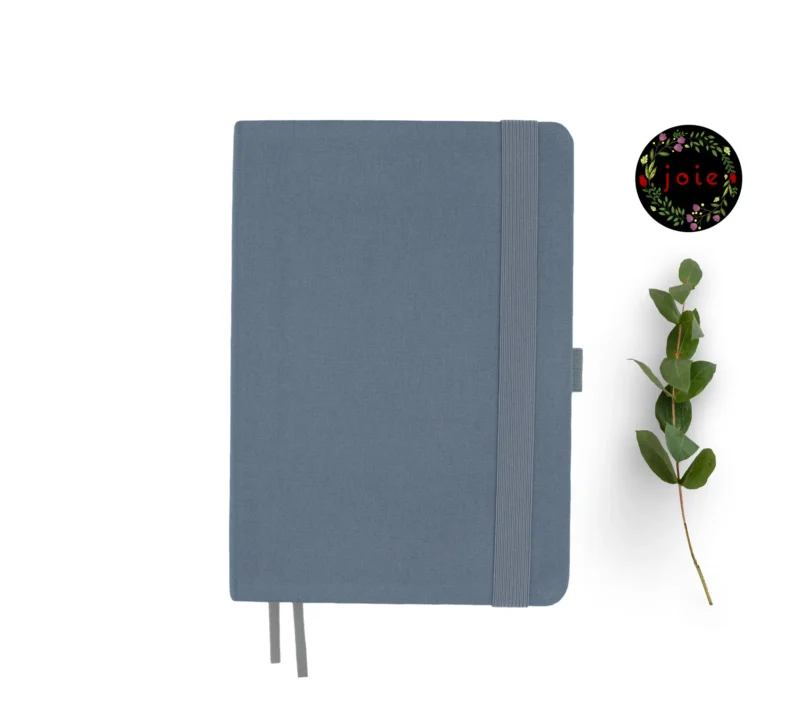 Gray fabric-covered notebook with rounded corners, matching elastic band, pen loop, and dual ribbon bookmarks, shown with a green leafy branch and a floral 'joie' logo